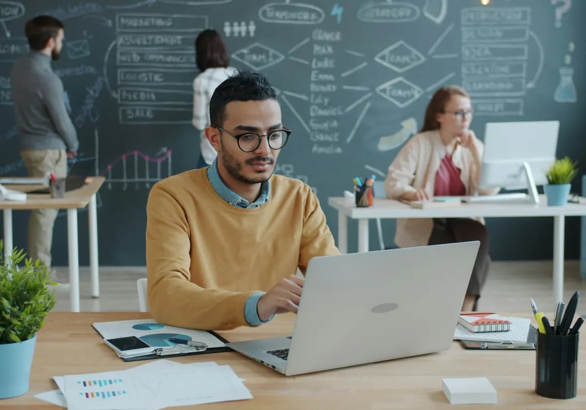 A startup founder working on a laptop while the team plans on a chalkboard